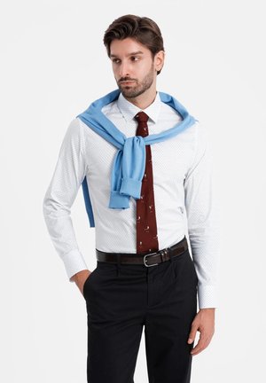 Camisa blanca con lunares azules, corbata burdeos con patrones azules y suéter azul claro atado alrededor de los hombros, combinado con pantalones oscuros.