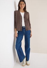 Brun stickad cardigan med knappar, bärs över en vit skjorta, ihop med blå utsvängda jeans och beige sneakers. Snyggt och avslappnat utseende.