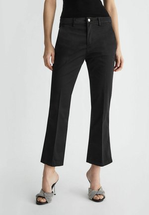 Black, high-waisted trousers with a straight leg design, featuring a front button closure and smooth texture. Worn with embellished high-heeled sandals.