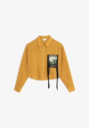 Mustard-colored cropped shirt with a textured finish, featuring a pocket holding a graphic print and black ribbon accents. Button-down design.
