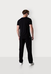 Schwarzes T-Shirt mit kurzen Ärmeln, kombiniert mit schwarzen, locker sitzenden Hosen. Dazu werden weiße Sneaker getragen; das T-Shirt hat ein kleines Logo auf dem Rücken.