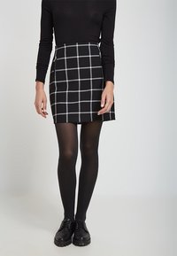 Falda negra con patrón de cuadrícula blanca, diseño ajustado, combinada con leggings negros y zapatos. Una blusa negra de manga larga completa el conjunto.