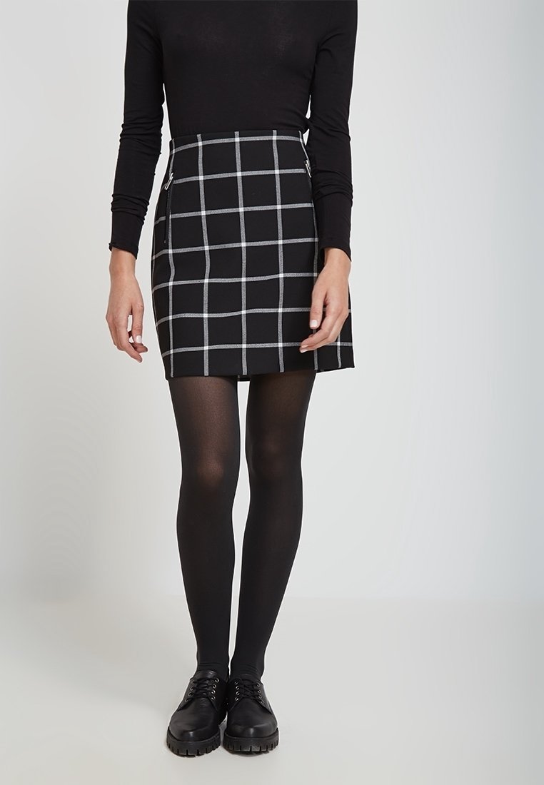 Falda negra con patrón de cuadrícula blanca, diseño ajustado, combinada con leggings negros y zapatos. Una blusa negra de manga larga completa el conjunto.