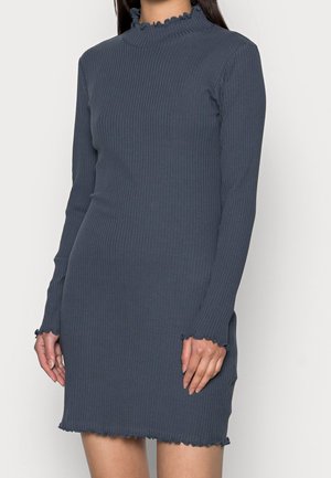 Robe longue ajustée en gris foncé côtelé avec des bords à volants au col, aux poignets et à l'ourlet, portée par une personne debout.