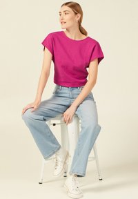 Fuchsia Crop-Top mit kurzen Ärmeln, kombiniert mit hellblauen Weitbeinen-Jeans und weißen Sneakern, sitzt auf einem Hocker. Einfacher Hintergrund.