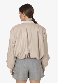 Beige faux leren bomberjack met verlaagde schouders, geribbelde kraag en elastische zoom, gedragen over grijs geruit shorts. Gladde textuur, minimalistisch ontwerp.