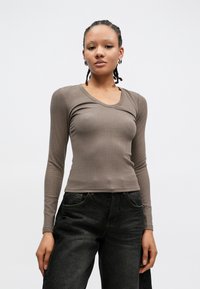 Hnědý top s dlouhým rukávem a úzkým střihem z žebrované látky s výstřihem do oblouku, v kombinaci s tmavě denimovými kalhotami s viditelnými detaily kapes.