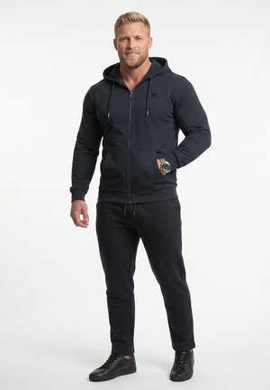 Blonder Mann trägt einen schwarzen Kapuzenpullover mit Reißverschluss, schwarze Hose, schwarze Sneaker und eine Armbanduhr und steht vor einem weißen Hintergrund.