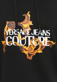 Črna kapuca z bogatimi zlatimi cvetličnimi vzorci in bold belim napisom "VERSACE JEANS COUTURE." Z vrvico z črnimi konci.