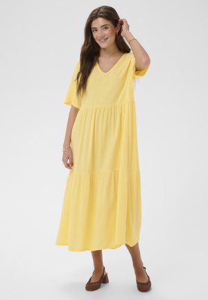 Mlada žena koja se smiješi, nosi široku žutu midi haljinu s V-izrezom i kratkim rukavima te smeđe pletene cipelice s blok petom, stoji na bijeloj pozadini.