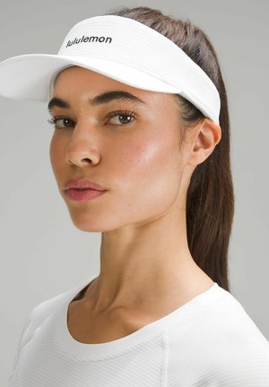 Visiera bianca Lululemon in tessuto strutturato, con visiera curva e logo ricamato. Indossata con una maglia bianca a maniche lunghe a coste.