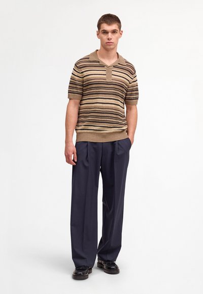Giovane uomo in piedi con le mani in tasca che indossa una polo a righe marroni a maniche corte, pantaloni larghi scuri e scarpe nere.
