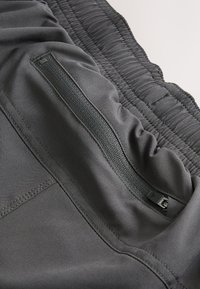 Pantalon gris foncé à taille élastique en gros plan, montrant une poche zippée avec des coutures détaillées sur un tissu lisse.