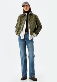 Chaqueta de color verde oliva con acolchado y cuello marrón, llevada sobre un suéter negro de cuello alto y una camisa blanca, combinada con jeans azules y zapatos marrones.