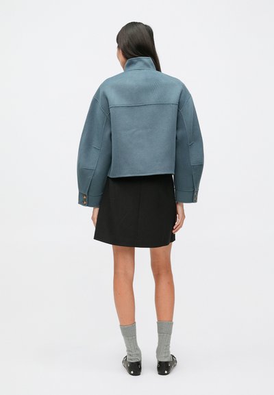 Niebieska, przycięta kurtka z wysokim kołnierzem, długimi rękawami i widocznymi szwami. Noszona na czarnej spódnicy, w połączeniu z szarymi prążkowanymi skarpetami i czarnymi butami.