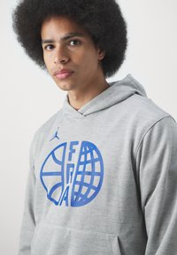 Felpa grigia con una grafica blu che combina un pallone da basket e un globo, con un piccolo logo sulla spalla. Tessuto morbido.