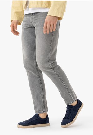 Muškarac u svijetlosivim trapericama, tamnoplavim tenisicama s bež đonovima, bijeloj košulji i bež jakni, stoji s jednom savijenom nogom.