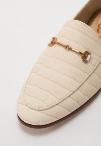 Beige loafers, kas izgatavoti no strukturēta sintētiska materiāla ar krokodila ādas rakstu. Priekšā ir zelta tonis papildus akcents.