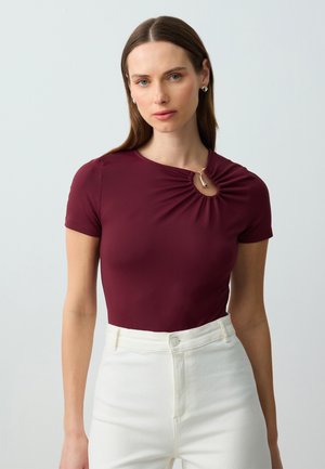 Top fitted borgogna con maniche corte, caratterizzato da un dettaglio arricciato e un accento dorato vicino al collo, abbinato a pantaloni bianchi a vita alta.