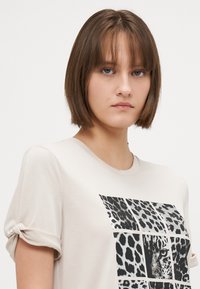 Beige tričko s krátkým rukávem, které na přední straně zdobí černý potisk leopardího vzoru v mřížkovém uspořádání. Měkká látka s volným střihem.