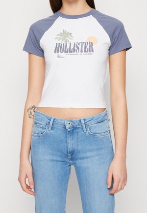 Weißes, kurzes T-Shirt mit blauen Raglanärmeln, mit dem Schriftzug "HOLLISTER", einer Palme und einem Sonnenmotiv. Kombiniert mit hellblauen Jeans.