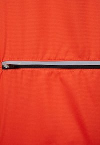 Una tela de color naranja brillante presenta un bolsillo horizontal con cremallera negra y detalles. La textura parece suave con un patrón en cuadrícula.