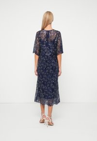 Robe en dentelle bleu marine avec des détails en sequins, manches courtes et longueur sous le genou. Elle présente une fermeture à lien au dos. Sandales à lanières.