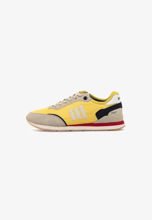 Sneaker giallo e beige con dettagli in suede, particolari neri e rossi, lacci e un audace logo "m" sul lato, visto dalla parte esterna.