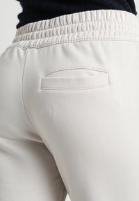 Ljust beige sweatshirtbyxor har en ribbad elastisk midja och en enda dekorativ bakficka med en rundad öppning. Slät tygtextur.