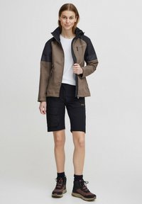 Waterdichte bruin en zwarte jas met ritszakken, gecombineerd met zwarte cargoshorts en stevige wandelschoenen. Gladde textuur en ritsafsluiting.