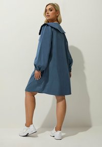 Vestido azul con cuello y mangas largas, de corte holgado, con un ribete negro con volantes y combinado con zapatillas deportivas blancas.
