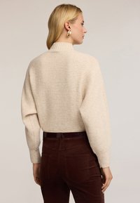 Beżowy sweter na drutach z wysokim kołnierzem i luźnym krojem, zestawiony z brązowymi sztruksowymi spodniami. Strój charakteryzuje się teksturowaną tkaniną i prostym wzorem.