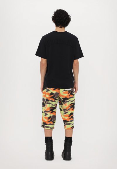 Svart t-skjorte med kort erm og logo på baksiden, parret med kne-lange camo-bukser i grønne, oransje, grå og svarte farger, samt svarte støvler.
