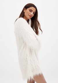 Gros gilet blanc duveteux en tissu texturé doux, avec des manches longues et une coupe ample, atteignant la mi-cuisse.
