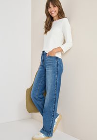 Witte lange mouwen top, blauwe high-waisted jeans en lichte sneakers. Outfit omvat een groene tote bag. Soepele texturen en een relaxte pasvorm.