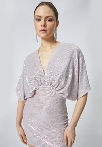 Robe en sequins rose avec un décolleté en V profond, des manches amples et une taille froncée. La texture lisse et les accents scintillants rehaussent le design.