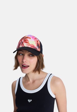 Femme portant un débardeur noir et blanc et une casquette avec un motif de palmier rose et orange, regardant sur le côté, la bouche légèrement ouverte.