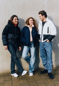 Tre persone stanno insieme, indossando diverse giacche: una felpa nera, un bomber blu navy e una felpa bianca con dettagli neri; tutti indossano pantaloni in denim.