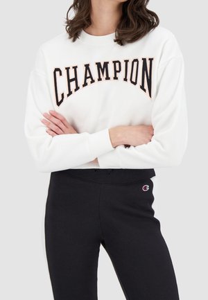 Persoon met een witte trui met de tekst "CHAMPION" en een zwarte broek met een klein Champion-logo, armen gekruist.
