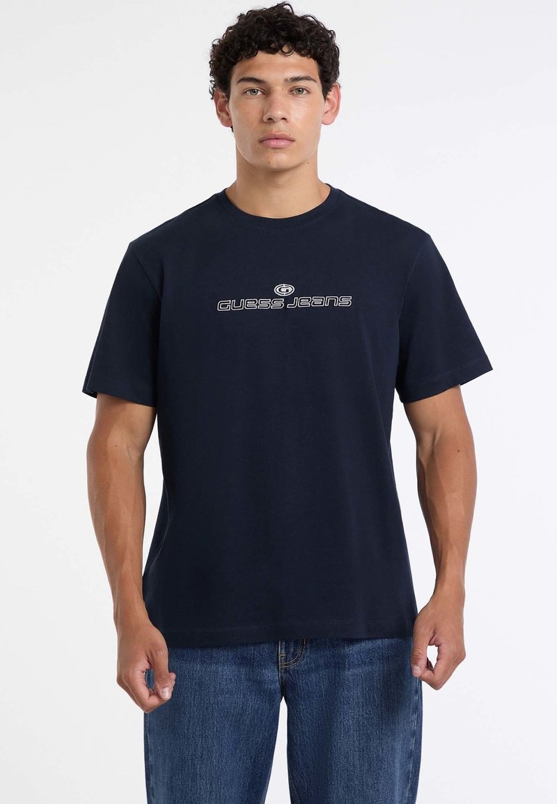 Tumesinine puuvillane T-särk lühikeste varrukatega, mille esiküljel on kontrastne valge "GUESS JEANS" logo. Kombineeritud siniste teksapükstega.