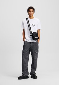 Sort crossbody taske med logo, hvid t-shirt med grafik, mørkegrå cargo bukser, sorte sko med tekstureret sål, stående foran en ensfarvet baggrund.