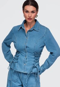 Femme portant une veste en jean bleu clair avec des côtés à lacets et un pantalon en jean assorti, debout les mains sur les hanches devant un fond uni.