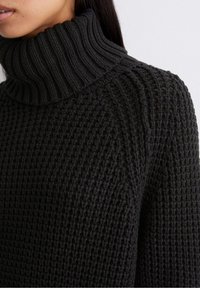 Pull en maille noire épaisse avec col roulé et motif texturé. La vue de côté montre sa forme distincte et son design confortable.