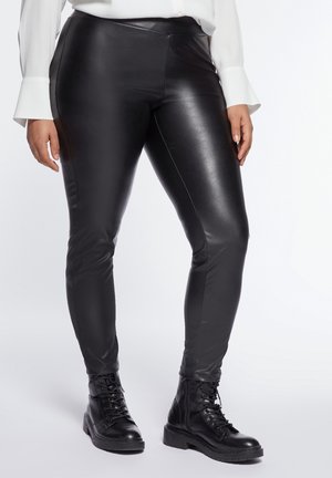 Henkilö pukeutuneena mustiin keinonahkaisiin leggingsseihin, mustiin nyörillisiin nilkkasaappaisiin ja valkoiseen pitkähihaiseen paitaan, jossa levenevät hihansuut.