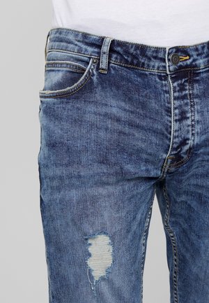 Niebieskie dżinsy z postrzępionym rozdarciem na górnej części uda, noszone z włożoną do środka białą koszulą na jednolitym jasnym tle.