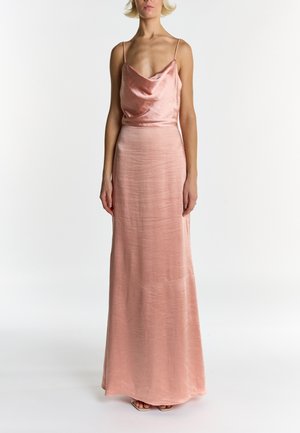 Donna che indossa un abito lungo fino al pavimento in raso color rosa cipria con sottoveste, spalline sottili e scollo a drappeggio, in piedi davanti a uno sfondo bianco uniforme.