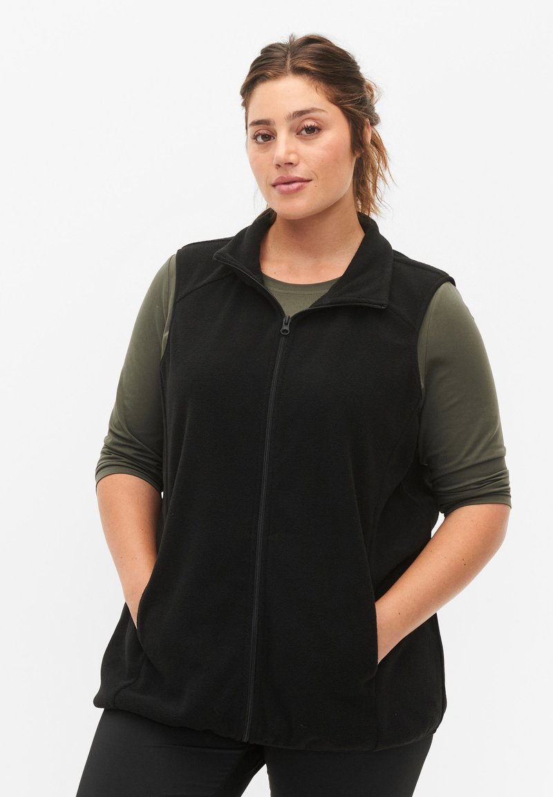 Gilet en polaire noir avec fermeture éclair devant, col montant et poches latérales, superposé à une chemise à manches longues verte, sur fond blanc.