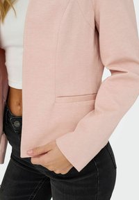 Femme portant un blazer rose clair sur un crop top blanc et un jean noir, tenant le bord avant du blazer d'une main.