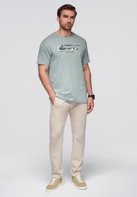 Lichtgroen T-shirt met een bedrukt graphic met de tekst "Simplicity in motion" gecombineerd met beige broek en beige sneakers. Glad stoffen textuur.