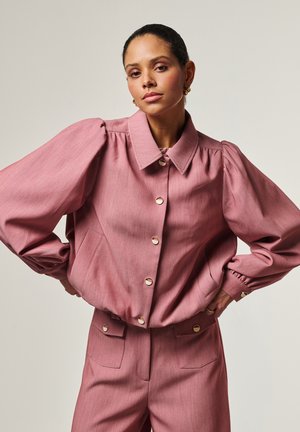 Vrouw met naar achteren gekamde haren, gekleed in een roze jas met knopen en pofmouwen, en bijpassende high-waisted broek, staand met handen in de zij.